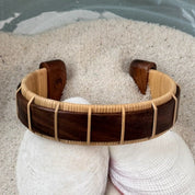 Rosewood Beam cuff bracelet resting on seashells and a smooth stone in sand, showing the bold open-weave cane design.