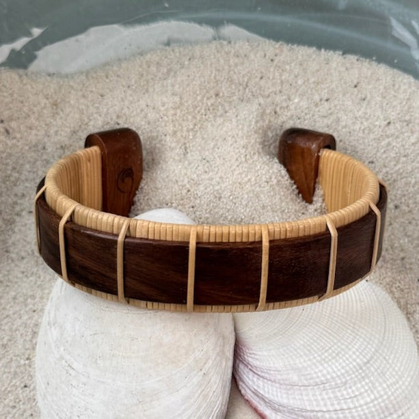 Rosewood Beam cuff bracelet resting on seashells and a smooth stone in sand, showing the bold open-weave cane design.