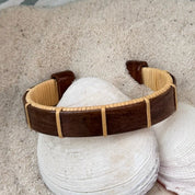 Rosewood Boardwalk cuff bracelet resting on seashells and a smooth stone in sand, showing the open-weave cane design and rich dark wood grain.
