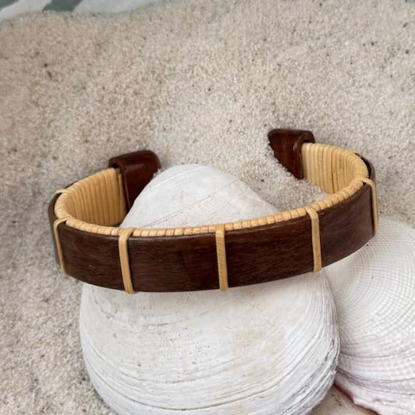 Rosewood Boardwalk cuff bracelet resting on seashells and a smooth stone in sand, showing the open-weave cane design and rich dark wood grain.