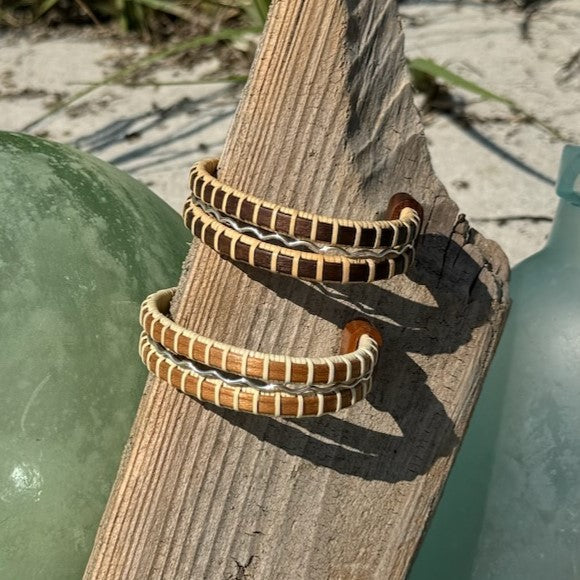 Handcrafted rosewood and cherry Waves triple cuff bracelets with sterling silver accents, resting on driftwood beside sea glass.