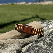 Handcrafted rosewood Vista cuff bracelet with a holly wood slider bead, resting on driftwood beside coiled rope.