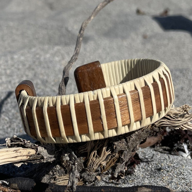 Handcrafted rosewood Legacy Cuff Bracelet resting on driftwood on a sandy beach with ocean waves in the background.