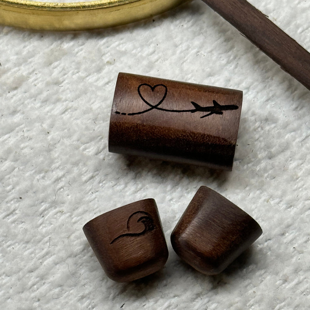 Close-up flat lay of the rosewood slider bead etched with an airplane and heart design alongside two rosewood end caps — one engraved with a wave-heart motif and one plain — showing the custom etching options for the Love In the Air Cuff Bracelet.