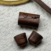 Close-up flat lay of the rosewood slider bead etched with an airplane and heart design alongside two rosewood end caps — one engraved with a wave-heart motif and one plain — showing the custom etching options for the Love In the Air Cuff Bracelet.
