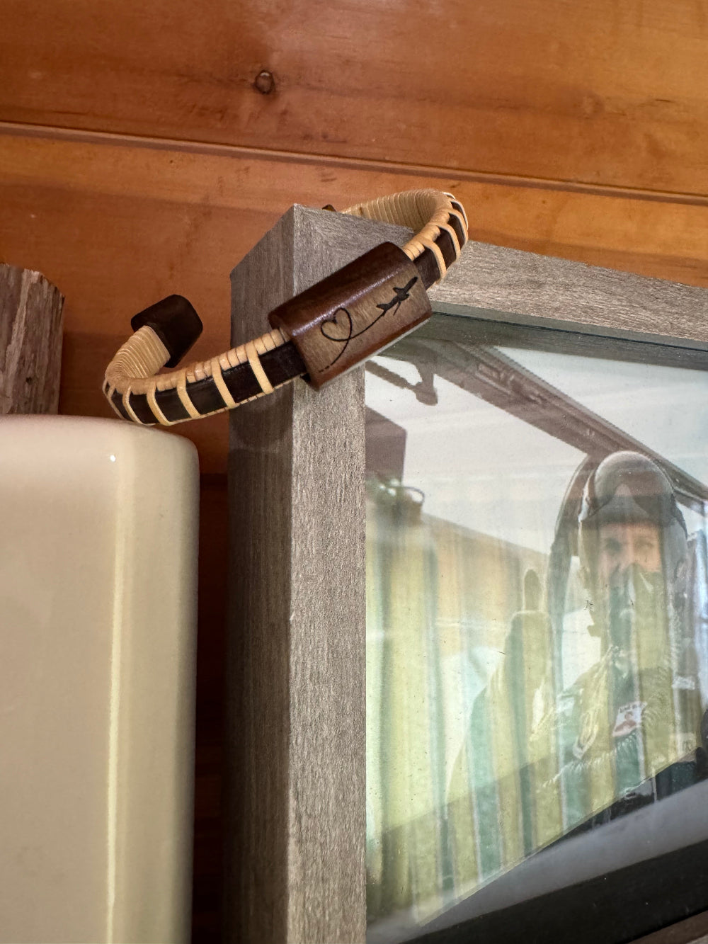 Close-up of the handcrafted rosewood Love In the Air Etched Cuff Bracelet showing the airplane and heart engraving on the rosewood slider bead, displayed on a picture frame with a pilot photo visible behind it.