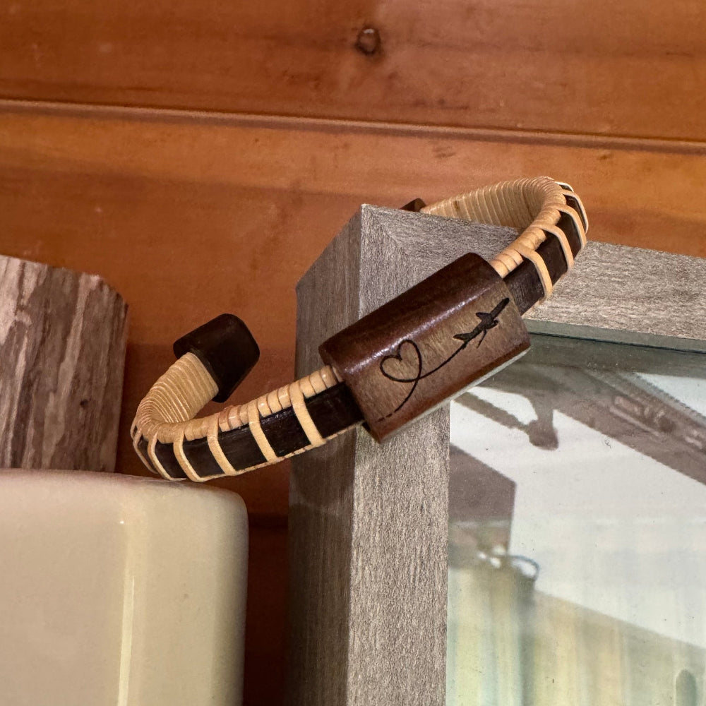 Handcrafted rosewood Love In the Air Etched Cuff Bracelet with a rosewood slider bead engraved with an airplane and heart design, balanced on a picture frame corner with a pilot photo in the background.