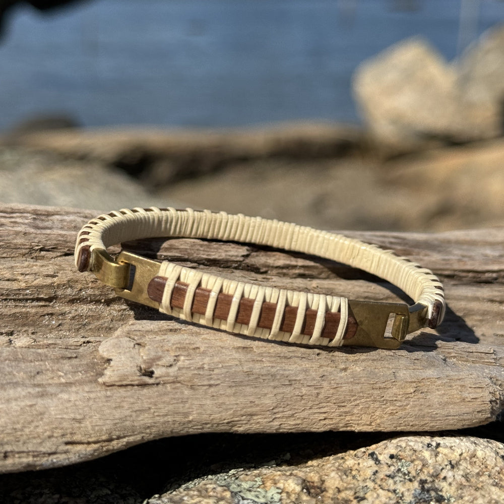 Handcrafted rosewood Nauti Clasp Bracelet with brass clasp resting on driftwood against coastal rocks with water in the background.