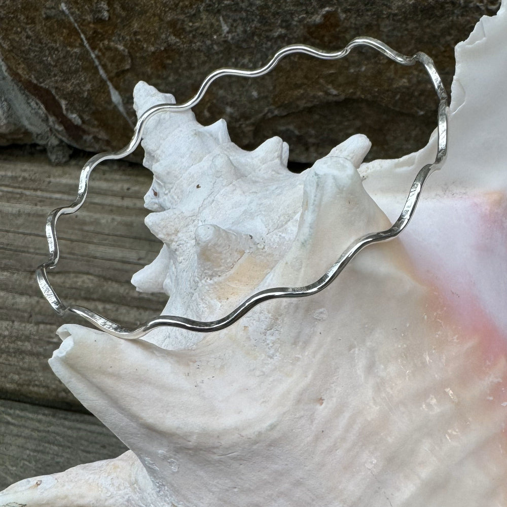 Close-up of a handcrafted hammered silver wave bangle bracelet draped over a conch shell, with rock and driftwood in the background.
