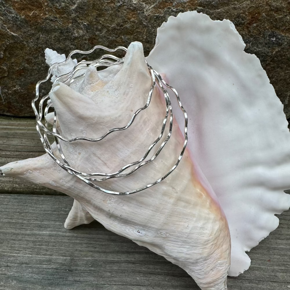 A stack of handcrafted hammered silver wave bangle bracelets draped over a conch shell, with rock and driftwood in the background.