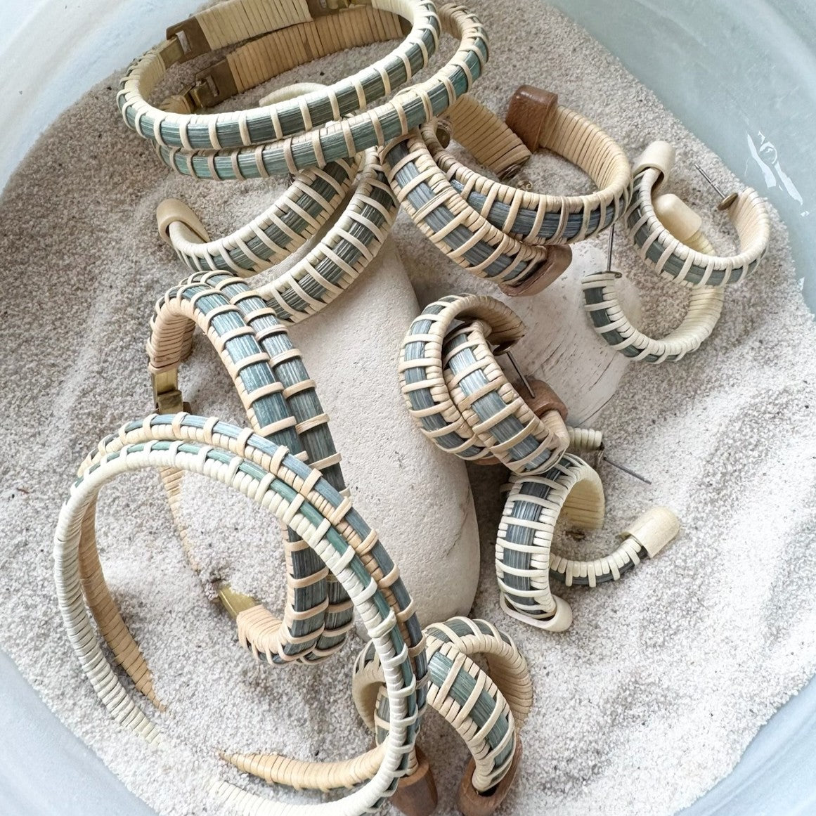 Handcrafted Summer Blues collection featuring woven bracelets, bangles, and earrings in Summer Sky blue cane, displayed in a bowl of sand.