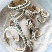 Handcrafted Summer Blues collection featuring woven bracelets, bangles, and earrings in Summer Sky blue cane, displayed in a bowl of sand.