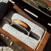 Handcrafted tungsten ring with cherry wood inlay resting on burlap lining inside an open engraved wooden gift box