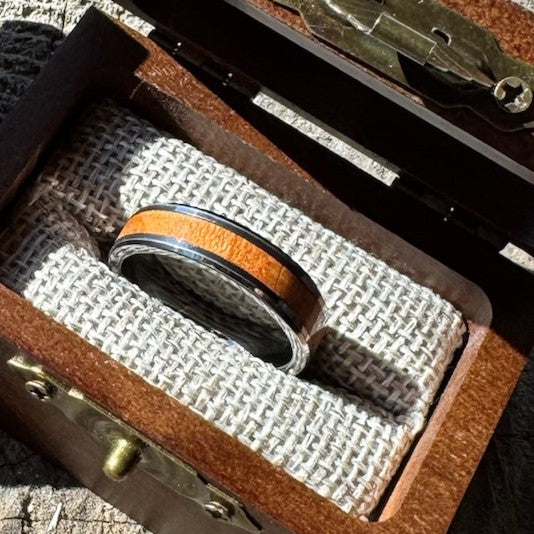 Handcrafted tungsten ring with cherry wood inlay resting on burlap lining inside an open engraved wooden gift box