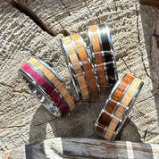 Four handcrafted 8mm tungsten double wood inlay Latitude rings in ebony cherry, rosewood white oak, purple heart white oak, and cherry white oak variants displayed on a wood stump, by Stonington Designs