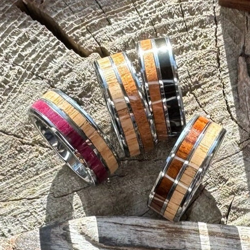 Four handcrafted 8mm tungsten double wood inlay Latitude rings in ebony cherry, rosewood white oak, purple heart white oak, and cherry white oak variants displayed on a wood stump, by Stonington Designs