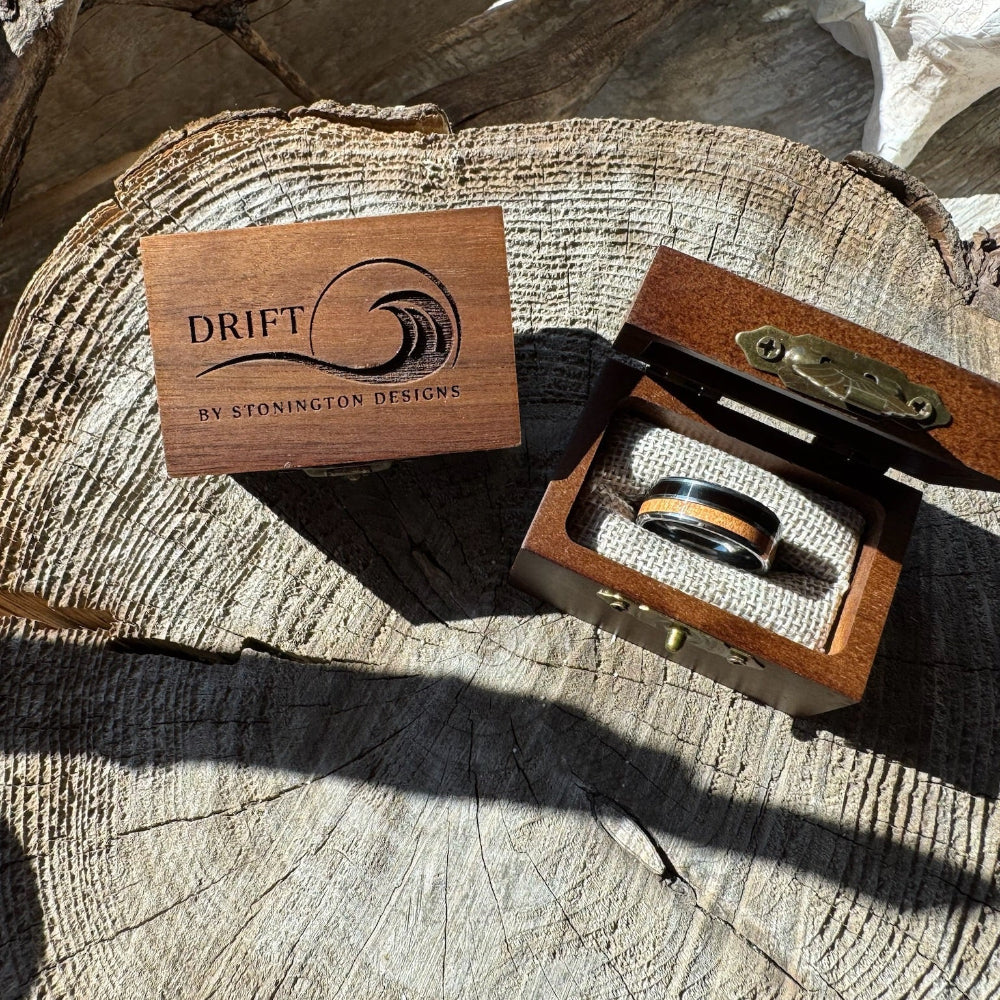 Handcrafted 8mm tungsten double wood inlay Latitude ring nestled in burlap inside an open engraved wooden gift box, displayed beside a closed Drift by Stonington Designs box on a wood stump.