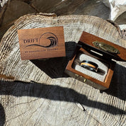 Handcrafted 8mm tungsten double wood inlay Latitude ring nestled in burlap inside an open engraved wooden gift box, displayed beside a closed Drift by Stonington Designs box on a wood stump.