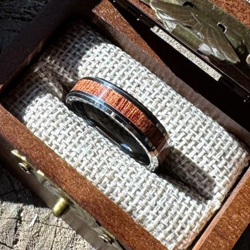 Handcrafted tungsten ring with mahogany wood inlay resting on burlap lining inside an open engraved wooden gift box