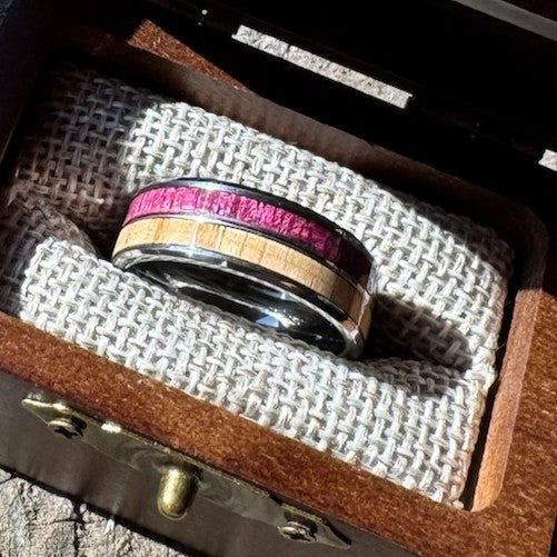 Handcrafted 8mm tungsten ring with purple heart and white oak double wood inlay resting on burlap lining inside an engraved wooden gift box.