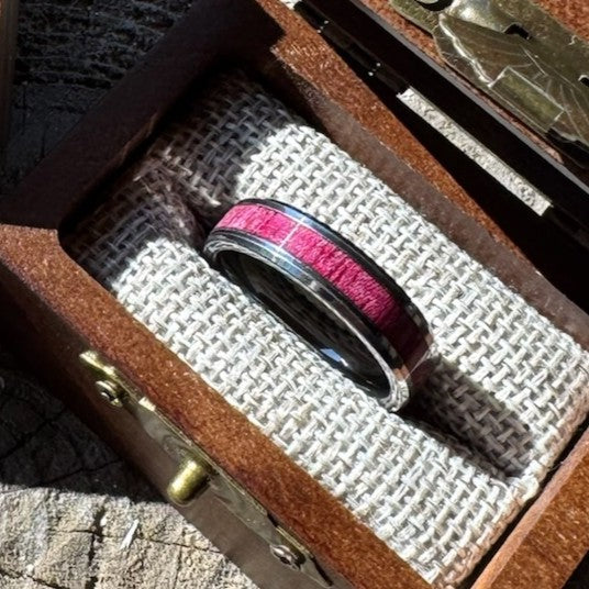 Handcrafted tungsten ring with purple heart wood inlay resting on burlap lining inside an open engraved wooden gift box