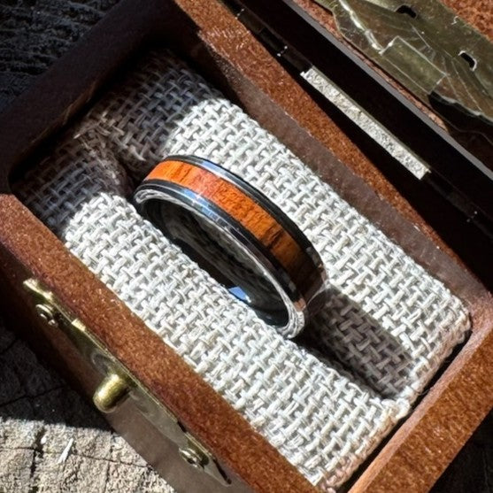 Handcrafted tungsten ring with rosewood inlay resting on burlap lining inside an open engraved wooden gift box