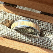 Handcrafted tungsten ring with white oak inlay resting in an open cherry wood commemorative box lined with burlap, part of the America250 Collection by Stonington Designs.