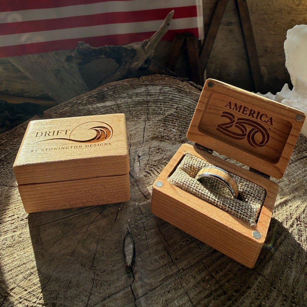 Two cherry wood commemorative boxes on a wood stump — one closed with the Drift by Stonington Designs logo, one open showing the America250 engraving inside the lid and a tungsten white oak ring nestled in burlap lining, with an American flag in the background.