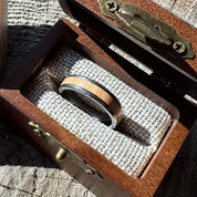 Handcrafted tungsten ring with white oak wood inlay resting on burlap lining inside an open engraved wooden gift box