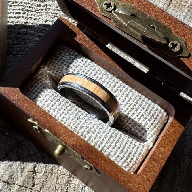 Handcrafted tungsten ring with white oak wood inlay resting on burlap lining inside an open engraved wooden gift box