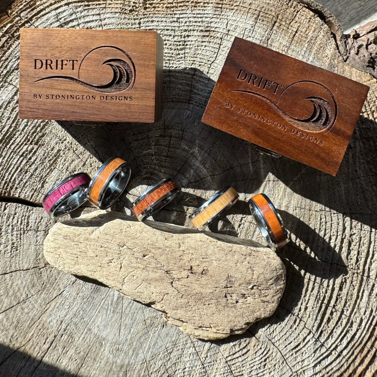 Four handcrafted tungsten wood inlay Heritage rings in purple heart, cherry, white oak, and rosewood resting on driftwood on a wood stump, with two engraved Drift by Stonington Designs cherry boxes behind them