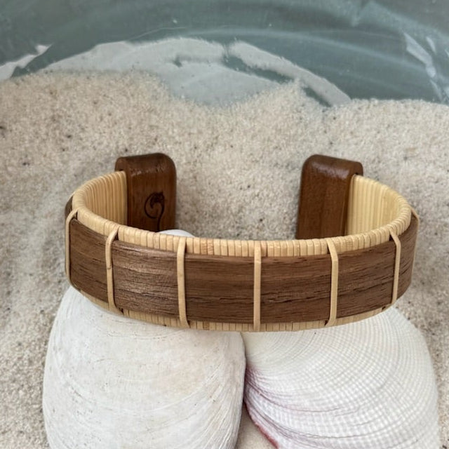 Walnut Beam cuff bracelet resting on seashells and a smooth stone in sand, showing the bold open-weave cane design.