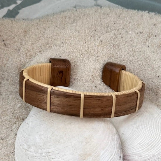 Walnut Boardwalk cuff bracelet resting on seashells and a smooth stone in sand, showing the open-weave cane design and deep walnut wood grain.
