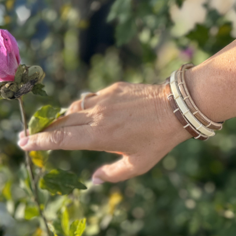 hree Breakers clasp bracelets in walnut, holly, and cherry stacked on a wrist outdoors with a pink flower and garden in the background.