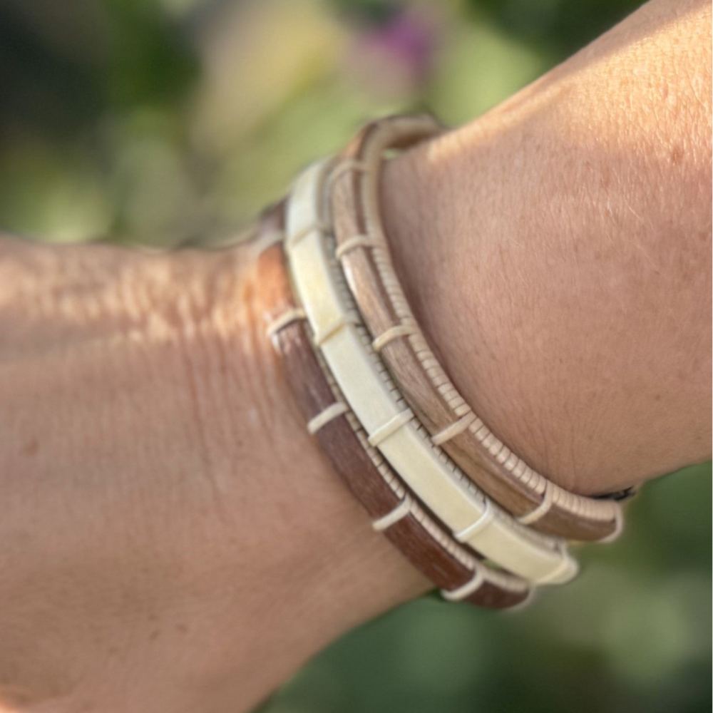Three Breakers clasp bracelets in walnut, holly, and cherry stacked on a wrist with a blurred garden background.