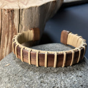 Handcrafted walnut Maverick Cuff Bracelet resting on a rock with a wood stump in the background, showing the deep warm walnut stave and holly weave construction.
