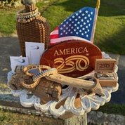 America 250 collection display outdoors featuring wood bracelets, a jewelry box with a ring in it, necklaces on display cards, an America 250 hitch cover, all in front of a woven bottle prop, and an American flag.