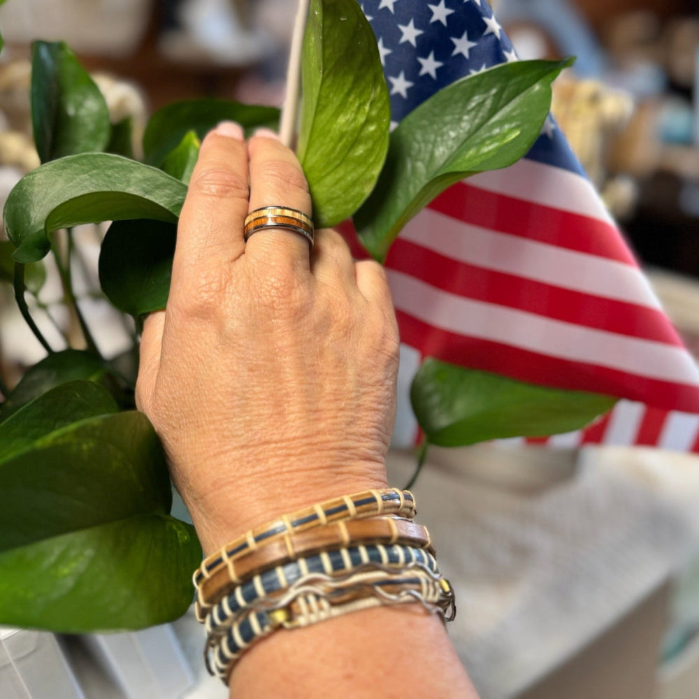 White oak and blue bamboo clasp bracelets stacked on a wrist, with Tungesten ring in finger, and an American flag and green leaves in the background