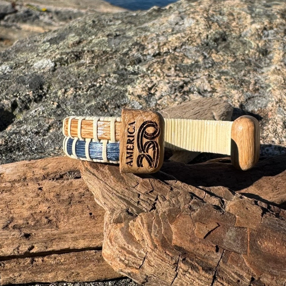 Close-up of the America 250 etched logo on the white oak endcap of a navy bamboo cuff bracelet, resting on driftwood.
