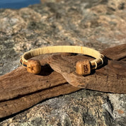 Handcrafted white oak and bamboo Patriot Cuff Bracelet resting on driftwood on coastal rocks, showing the star-etched end cap on one side and the America250 logo end cap on the other, part of the licensed America250 Collection by Stonington Designs.