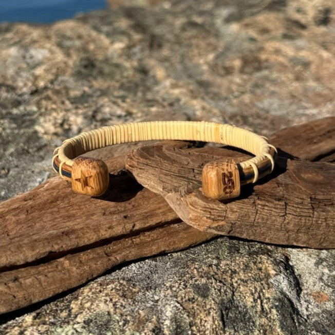 Handcrafted white oak and bamboo Patriot Cuff Bracelet resting on driftwood on coastal rocks, showing the star-etched end cap on one side and the America250 logo end cap on the other, part of the licensed America250 Collection by Stonington Designs.