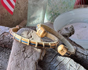 Two handcrafted white oak and navy bamboo Patriot Cuff Bracelets displayed on driftwood with an American flag tote in the background, showing the star and America250 etched white oak end caps, part of the licensed America250 Collection by Stonington Designs.