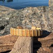 Handcrafted white oak Maverick America250 Cuff Bracelet balanced on driftwood against coastal rocks with water in the background, showing the premium upcycled white oak stave and handwoven construction made by military spouse artisans at Stonington Designs.