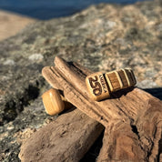 Close-up of the handcrafted white oak Maverick America250 Cuff Bracelet resting on driftwood and coastal rock, showing the America250 logo etched on the white oak end cap, part of the licensed America250 Collection by Stonington Designs.