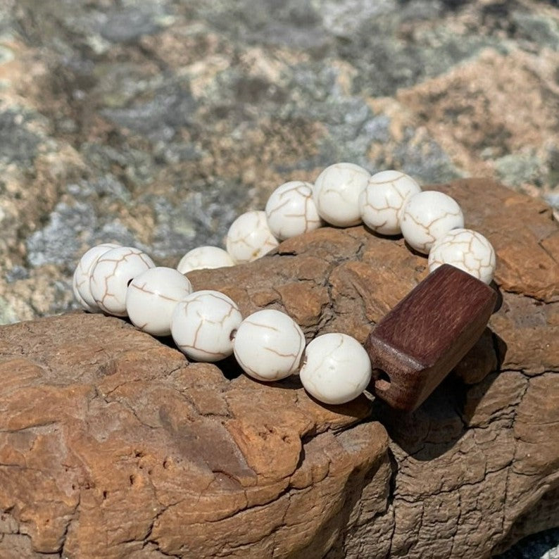 Beachcomber bracelet with white turquoise stones and a rectangular wood bead resting on driftwood and granite rocks.