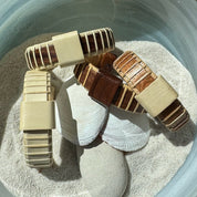 Four handcrafted Island Cuff Bracelets in holly, cherry, and rosewood variants with holly and rosewood slider beads, arranged in sand with a seashell and smooth stone, made by military spouse artisans at Stonington Designs