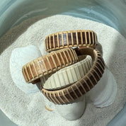 Four handcrafted Legacy Cuff Bracelets in walnut, rosewood, cherry, and holly variants arranged on seashells and sand, made by military spouse artisans at Stonington Designs.