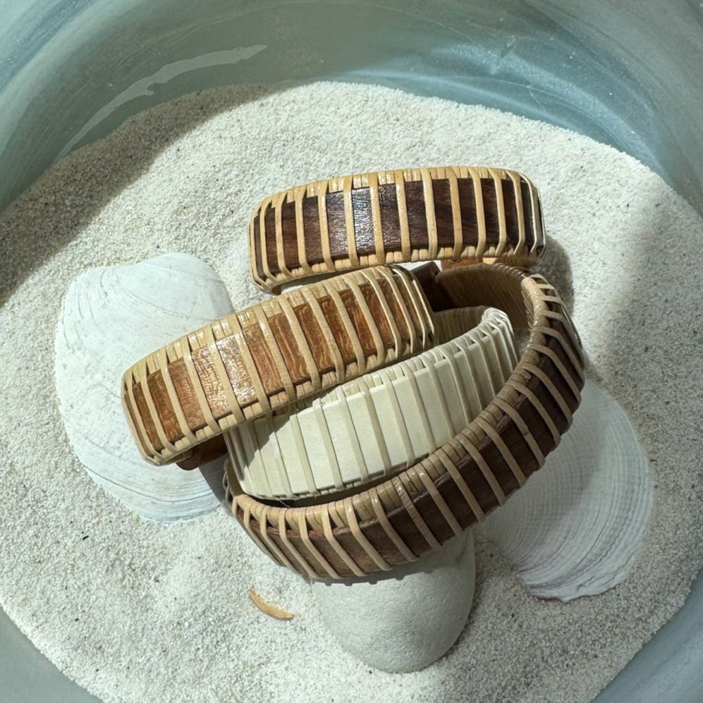 Four handcrafted Legacy Cuff Bracelets in walnut, rosewood, cherry, and holly variants arranged on seashells and sand, made by military spouse artisans at Stonington Designs.