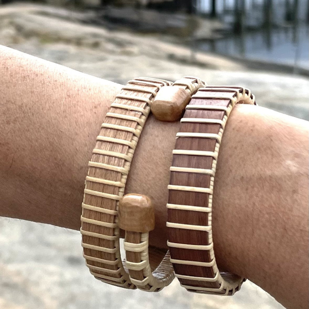 Two handcrafted Maverick Cuff Bracelets in white oak and rosewood worn stacked on a wrist with a coastal waterway in the background, showing the bracelet's versatility for stacking.