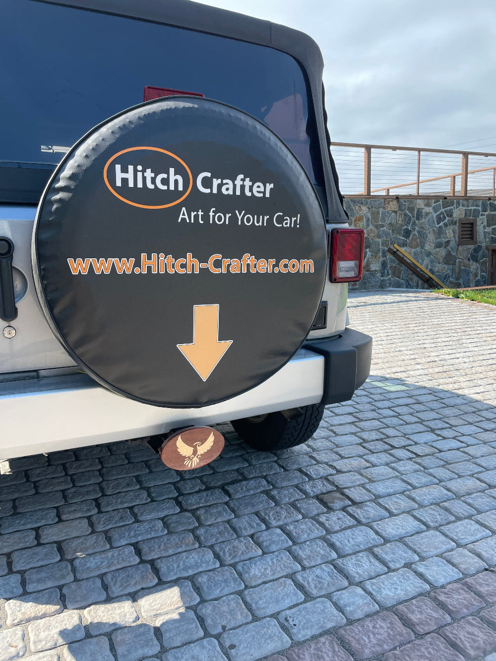 hitch crafter logo with arrow on Jeep wheel cover with arrow pointing to wooden eagle hitch cover.